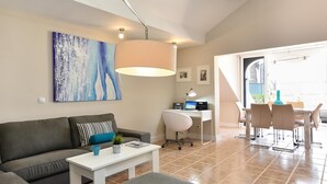 A modern living room with a grey sofa, a white coffee table, and a desk with a computer. There is a large abstract painting on the wall and a dining area with chairs and a table.