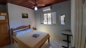 Un dormitorio con techo de madera, una cama con cubrecama color beige, una mesita de noche, un escritorio pequeño y una ventana con contraventanas.
