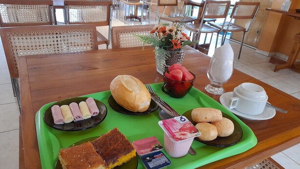 Bandeja de desayuno con pasteles, pan y un bol de fruta sobre una mesa en un comedor con sillas y mesas de mimbre.