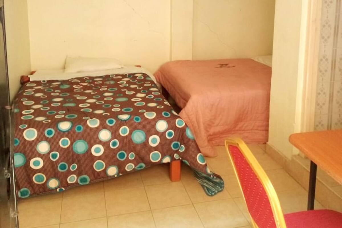 A small, sparsely furnished room with two beds, one with a polka dot bedspread and the other with a solid color. There is a single chair and a small table.