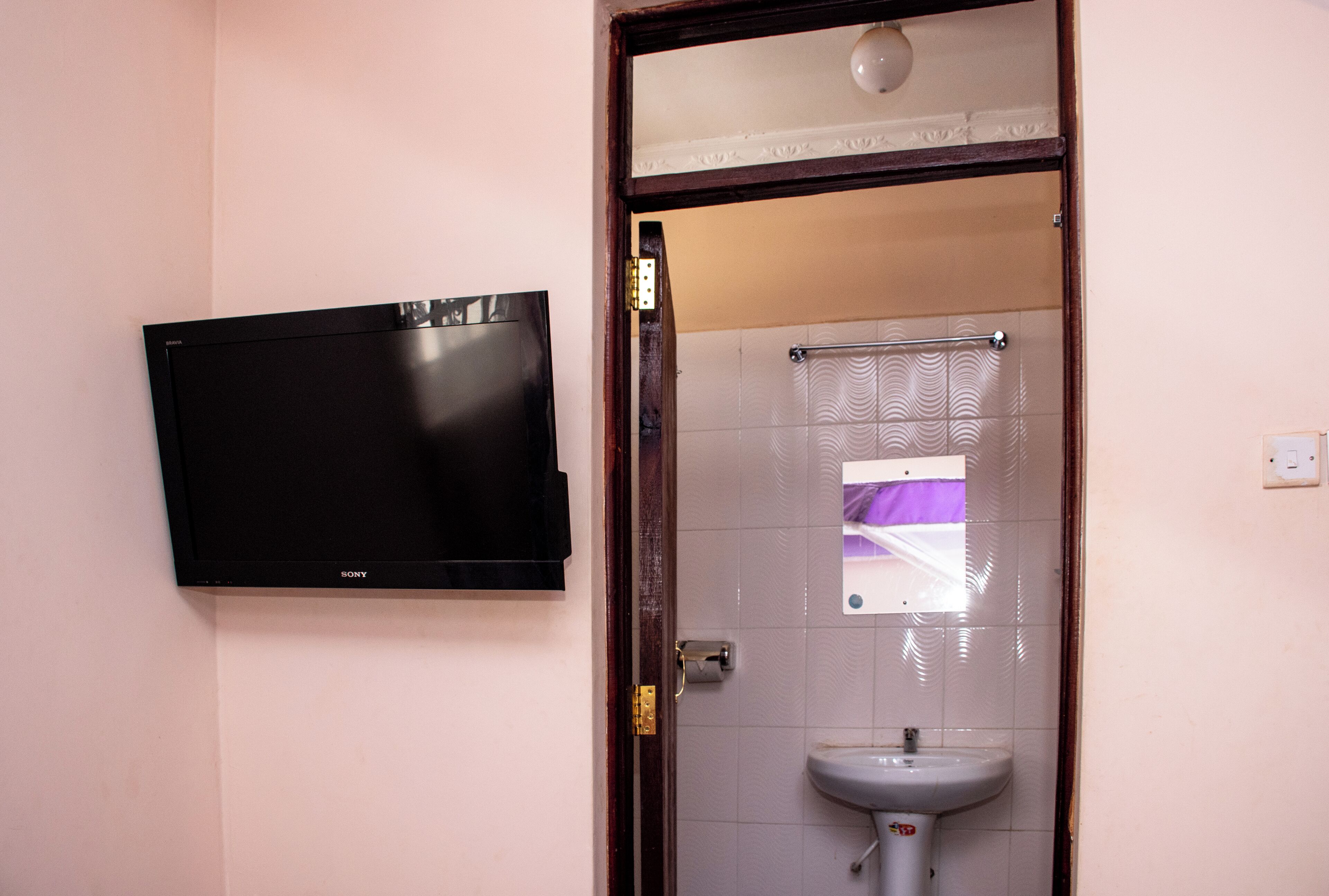 A bathroom with a black flat-screen TV mounted on the wall, a white sink, and a shower with a towel rack.