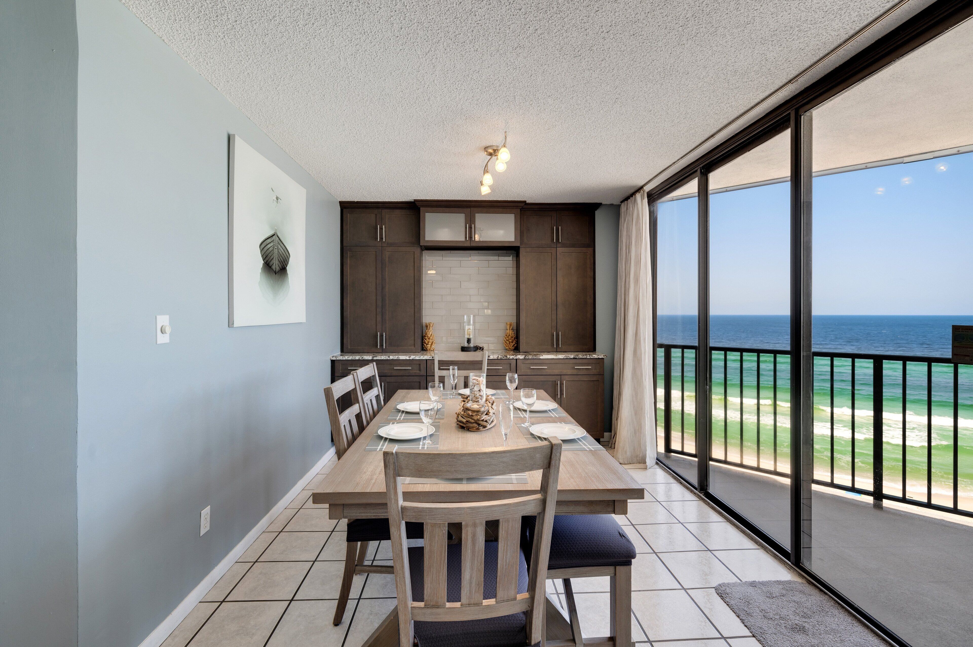 A dining area with a table set for four, a kitchen with dark cabinets and a white backsplash, and a large sliding glass door leading to a balcony with a view of the ocean.