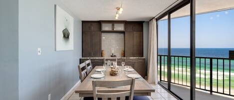A dining area with a table set for four, a kitchen with dark cabinets and a white backsplash, and a large sliding glass door leading to a balcony with a view of the ocean.
