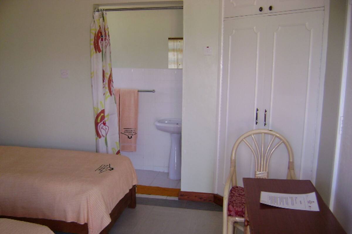 A bedroom with two beds, a wooden chair, a small table with a piece of paper, a bathroom with a sink and toilet, and a door.
