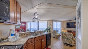 Una cocina con encimeras de granito, armarios de madera, lavavajillas y un tapete rojo. Hay una zona de comedor con mesa y sillas, un ventanal con vistas y un televisor.