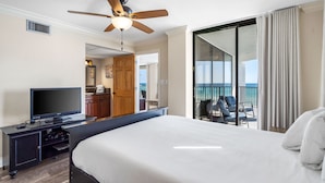 A hotel room with a bed, a TV, a ceiling fan, a balcony with chairs, and a view of the ocean.