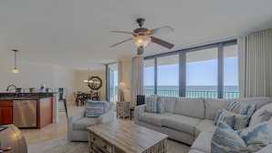 A spacious living area with a large sofa, a coffee table, and a ceiling fan. There is a kitchen area with a dishwasher and a dining table with chairs. The room has large windows offering a view of the ocean.