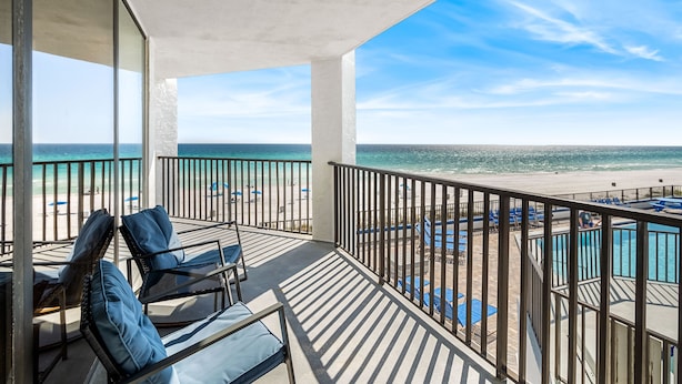 A balcony with two lounge chairs, a pool area with blue lounge chairs, and a view of the beach and ocean.