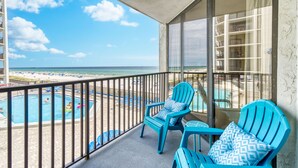 Un balcon avec vue sur une plage et une piscine, agrémenté de chaises bleues et d’une rambarde en verre.