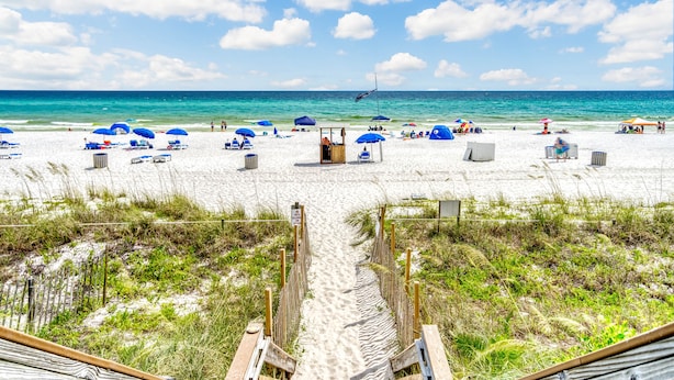 Une plage avec un sentier de sable menant à l’eau, des parasols bleus et des chaises de plage.