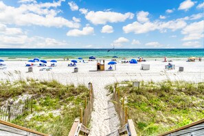 A beach with a sandy path leading to the water, blue umbrellas, and beach chairs.