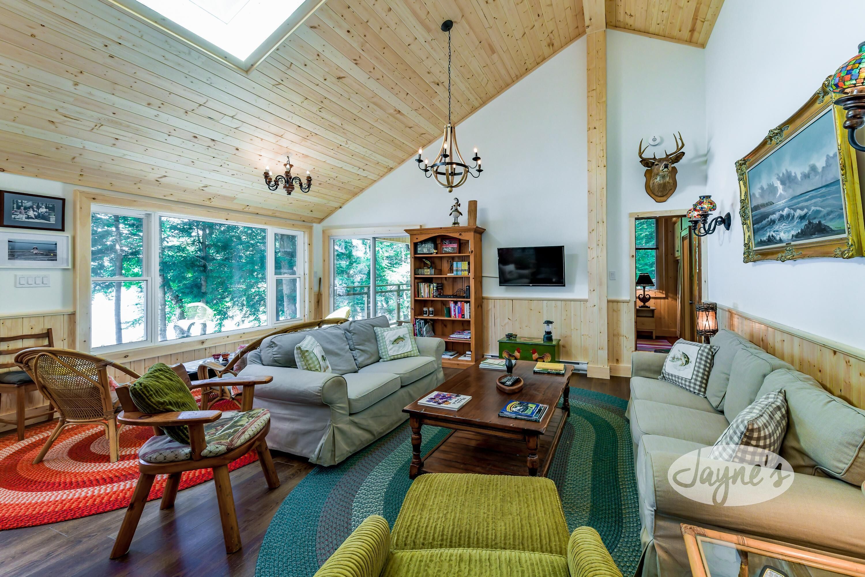 A spacious living room with a high ceiling, wooden beams, a chandelier, a fireplace, and a large window offering a view of trees.