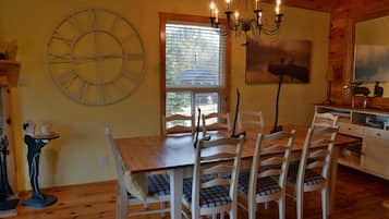 A dining area with a wooden table and chairs, a large wall clock, and a chandelier.