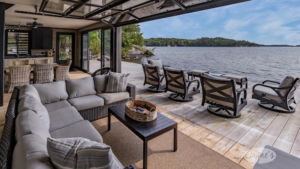 A modern outdoor living area with a sofa, chairs, and a table, overlooking a lake with a forested island in the distance. - Ultimate Boathouse Entertaining, New 6 bdrm, 8 bath Cottage/Boathouse! Hot tub! (Port Carling)