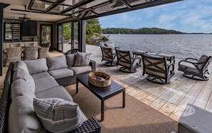 A modern outdoor living area with a sofa, chairs, and a table, overlooking a lake with a forested island in the distance. - Ultimate Boathouse Entertaining, New 6 bdrm, 8 bath Cottage/Boathouse! Hot tub! (Port Carling)