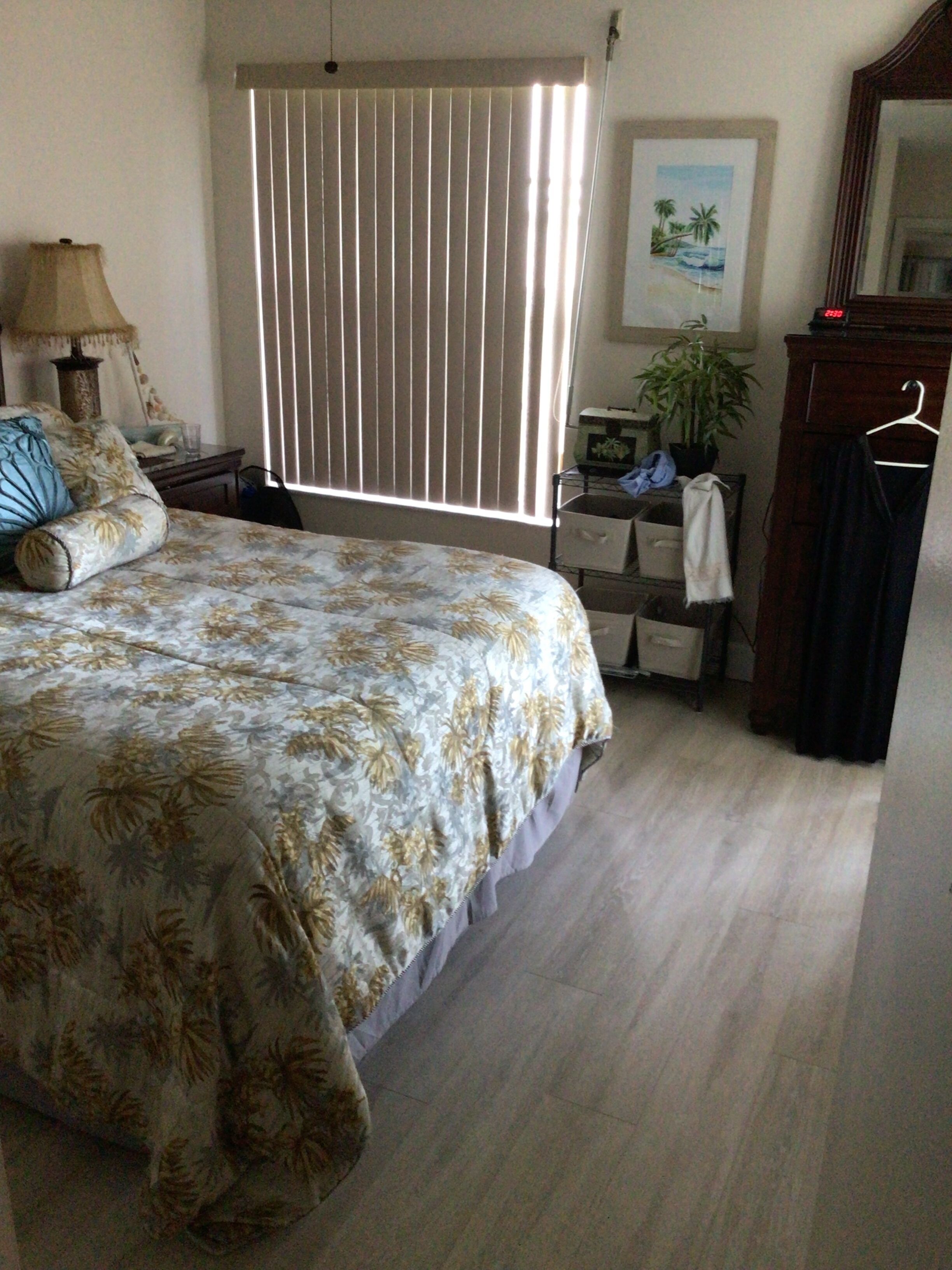 Une chambre à coucher comprenant un lit, une table de chevet, une commode, une fenêtre avec des stores, un tableau encadré accroché au mur et une plante posée sur la commode.