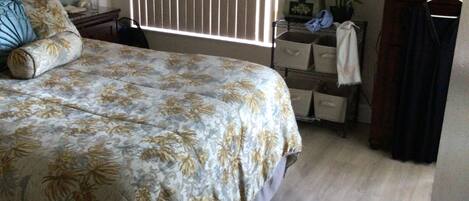 Une chambre à coucher comprenant un lit, une table de chevet, une commode, une fenêtre avec des stores, un tableau encadré accroché au mur et une plante posée sur la commode.