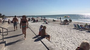 En strand med folk som promenerar och sitter på sanden, parasoller och en klar himmel.