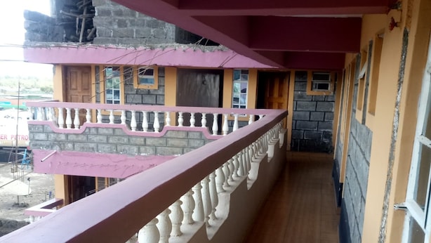 A corridor with a balcony railing, a stone wall, and a yellow building with a red roof. - 2030 Guest House (Nakuru)
