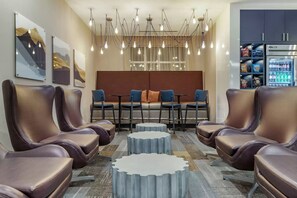 A modern hotel lobby with leather chairs, a central dining table with blue chairs, a patterned carpet, and a refrigerator stocked with beverages. - DoubleTree by Hilton Greenville Woodruff Road (Greenville)