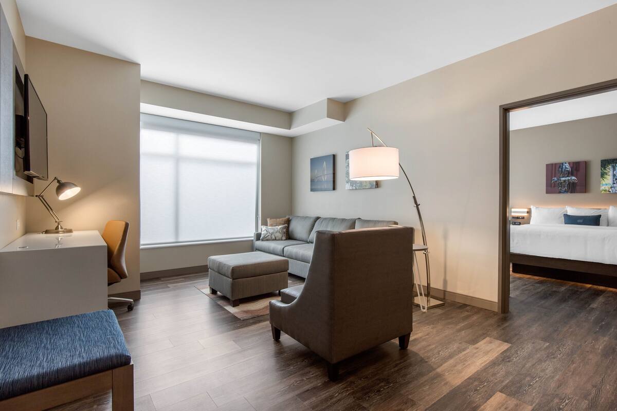 Een moderne woonkamer met een bank, poef en fauteuil. Er is een bureau met een lamp, een groot raam en kunst aan de muur.