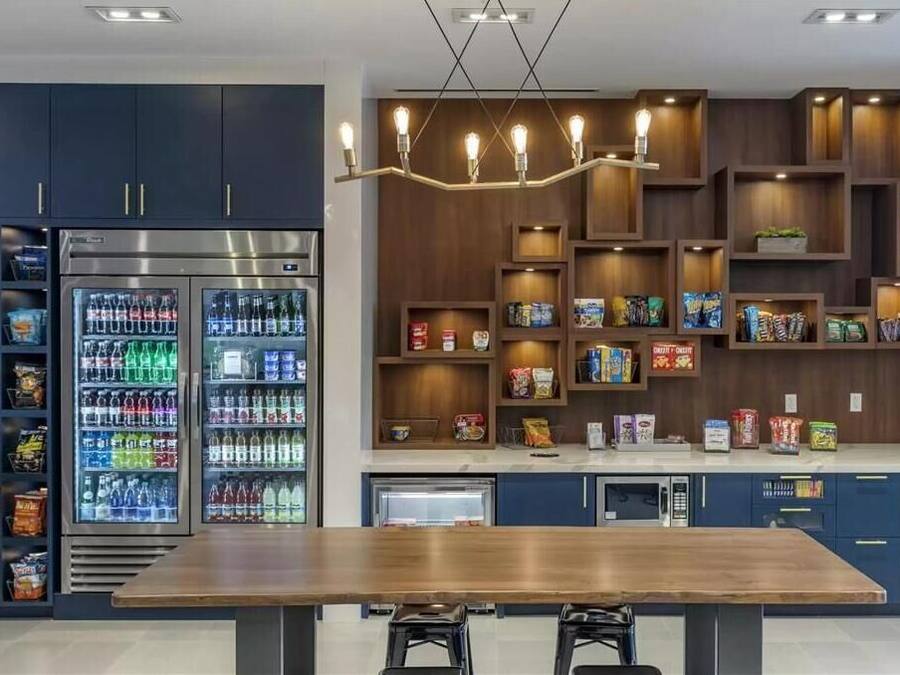 Uma cozinha moderna com mesa de madeira, geladeiras embutidas e prateleiras com lanches e bebidas.