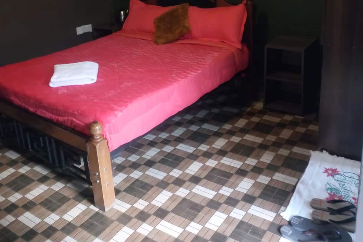 A bed with a red bedspread and pillows, a checkered floor, and a pair of slippers on a mat.