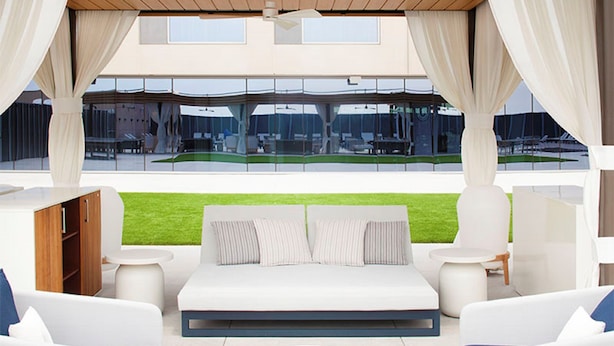 Un espace extérieur moderne aménagé avec un canapé et des fauteuils, une petite table et une vue sur un terrain de sport grâce à de grandes fenêtres.