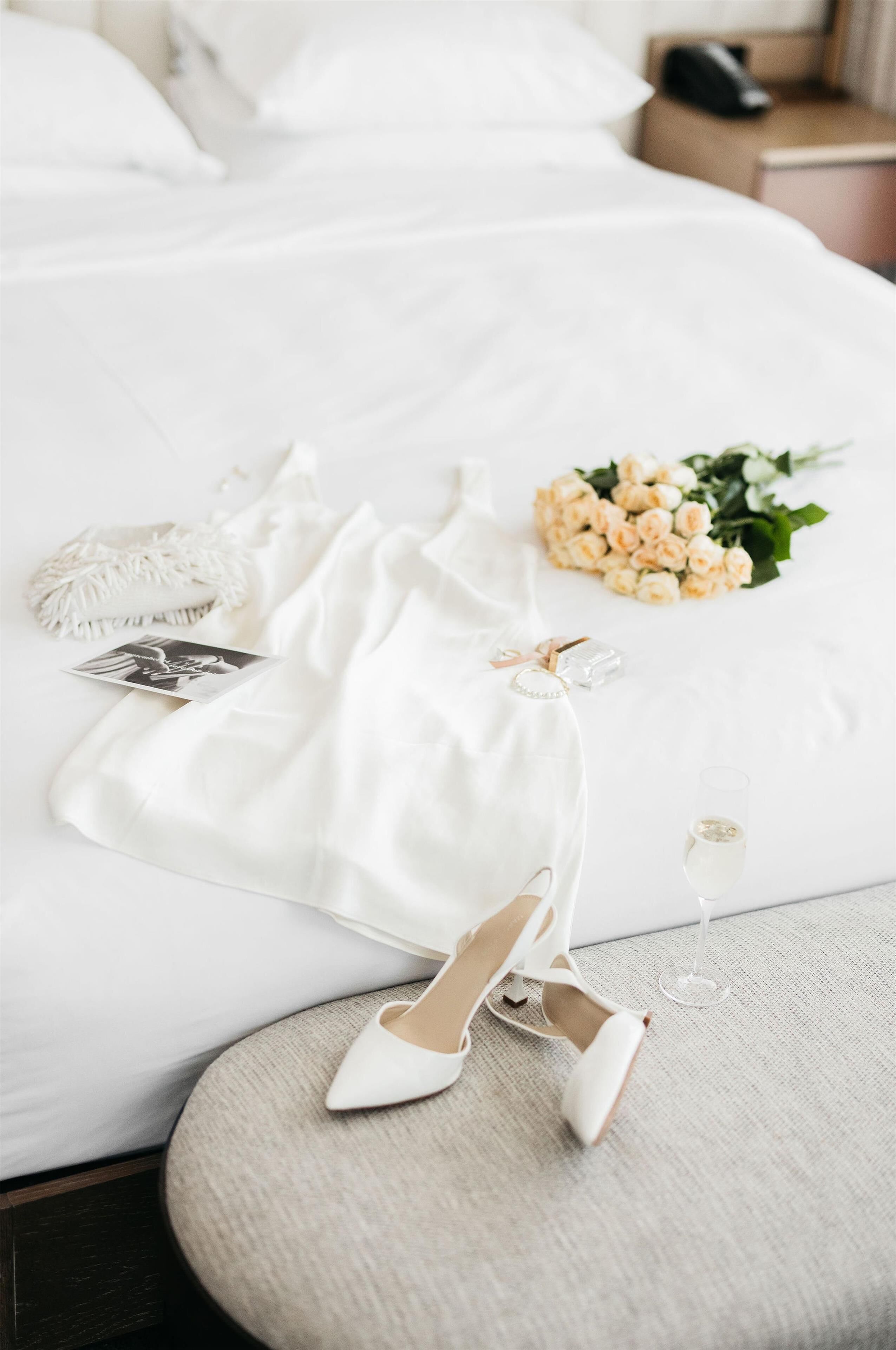 A bed with white bedding, a bouquet of flowers, a glass of champagne, and a pair of white shoes.