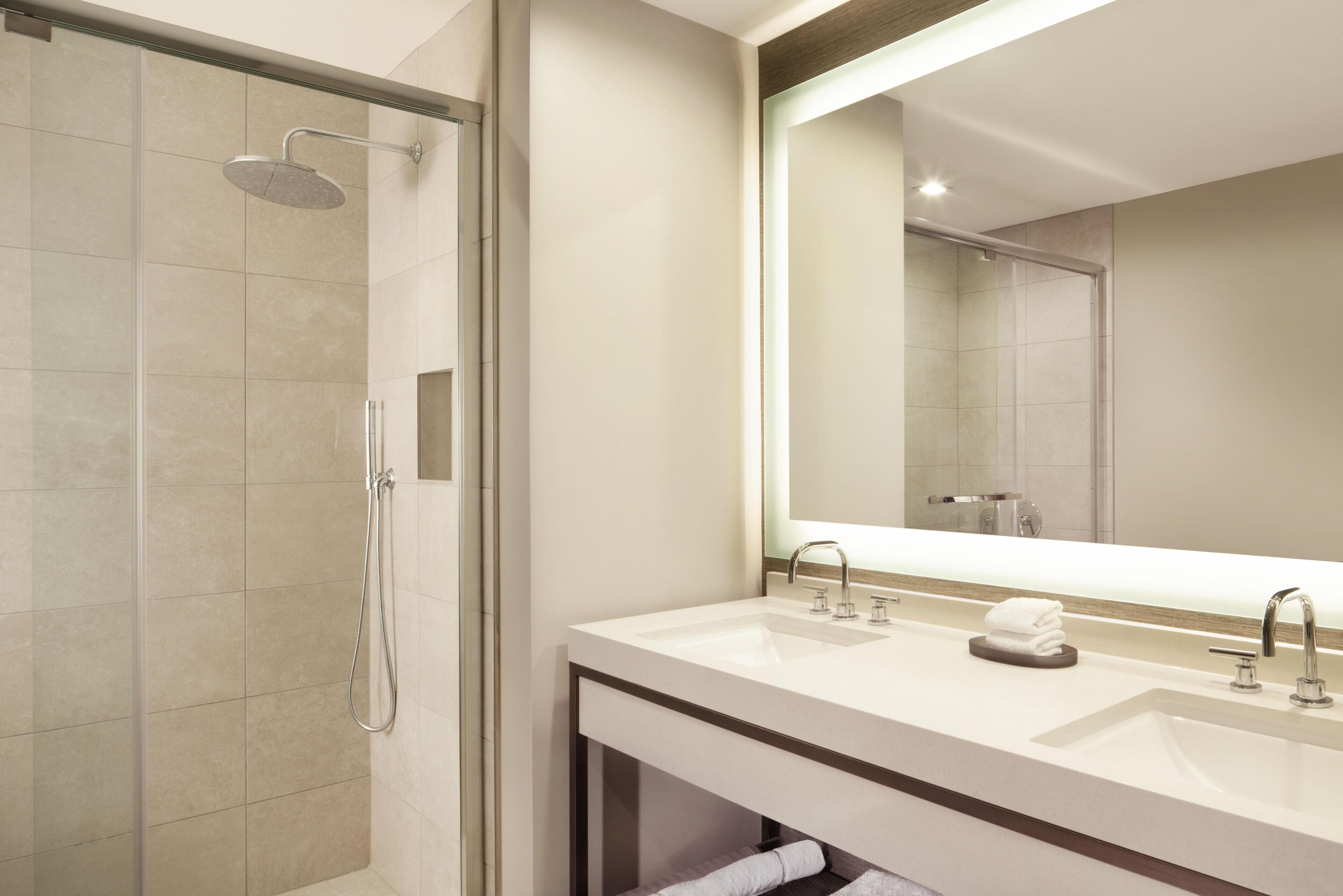 Un baño moderno con ducha de vidrio, un espejo grande y dos lavamanos.