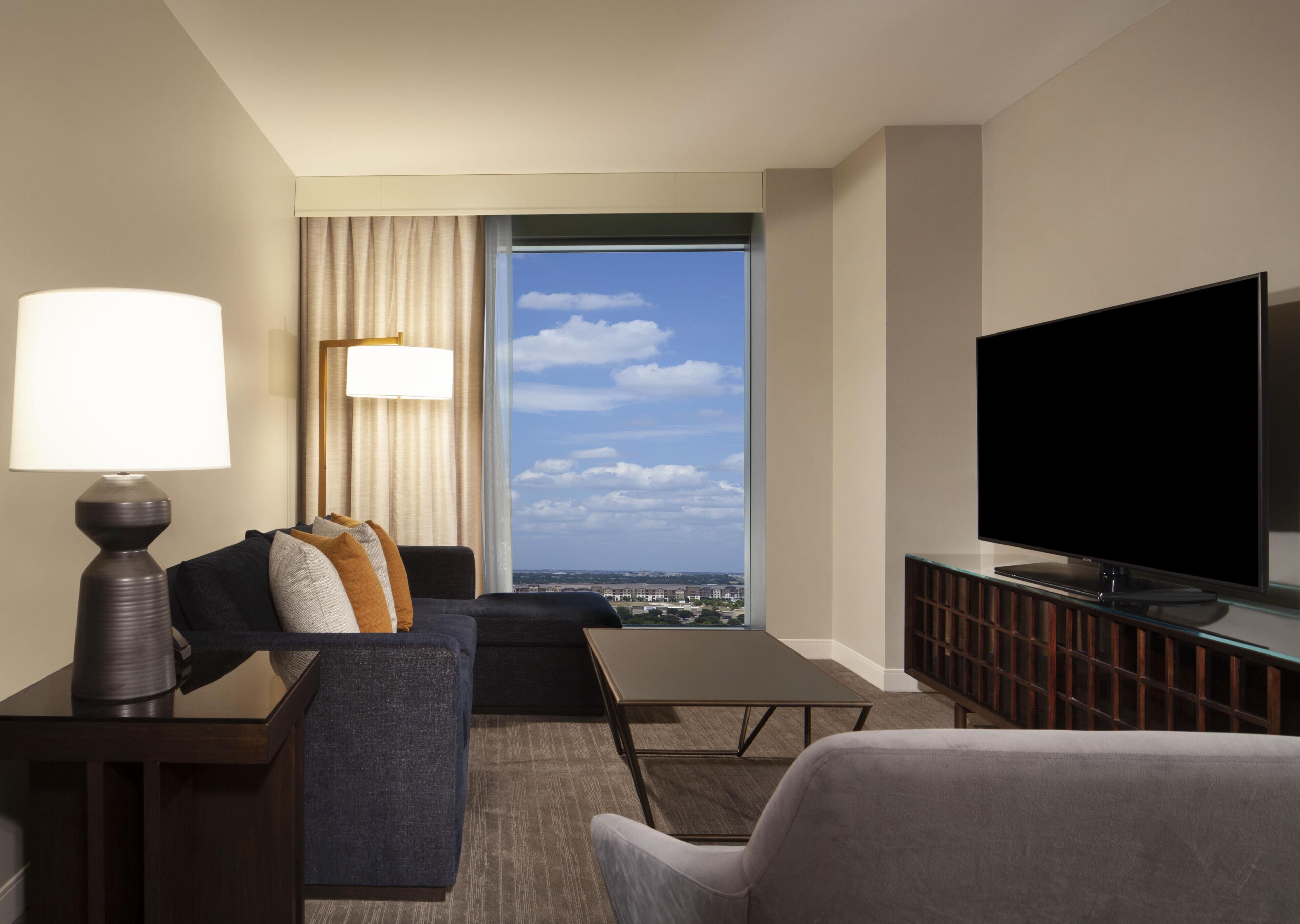 Una habitación de hotel con un ventanal grande, un televisor de pantalla plana, un sofá, una mesa y una lámpara.