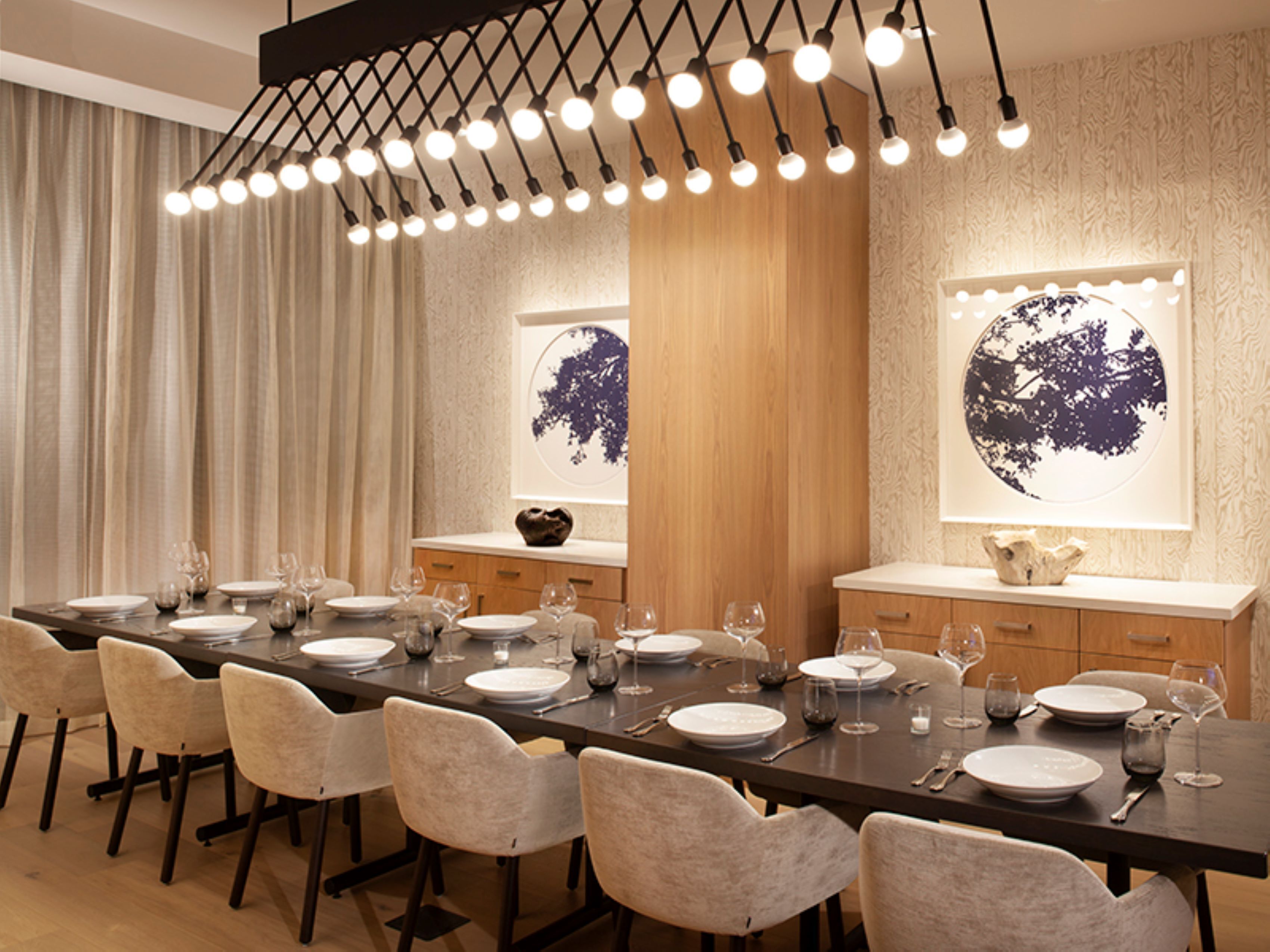 Un espace de salle à manger moderne, avec une longue table dressée pour un repas, agrémenté d’un lustre, de boiseries et d’œuvres d’art aux murs.