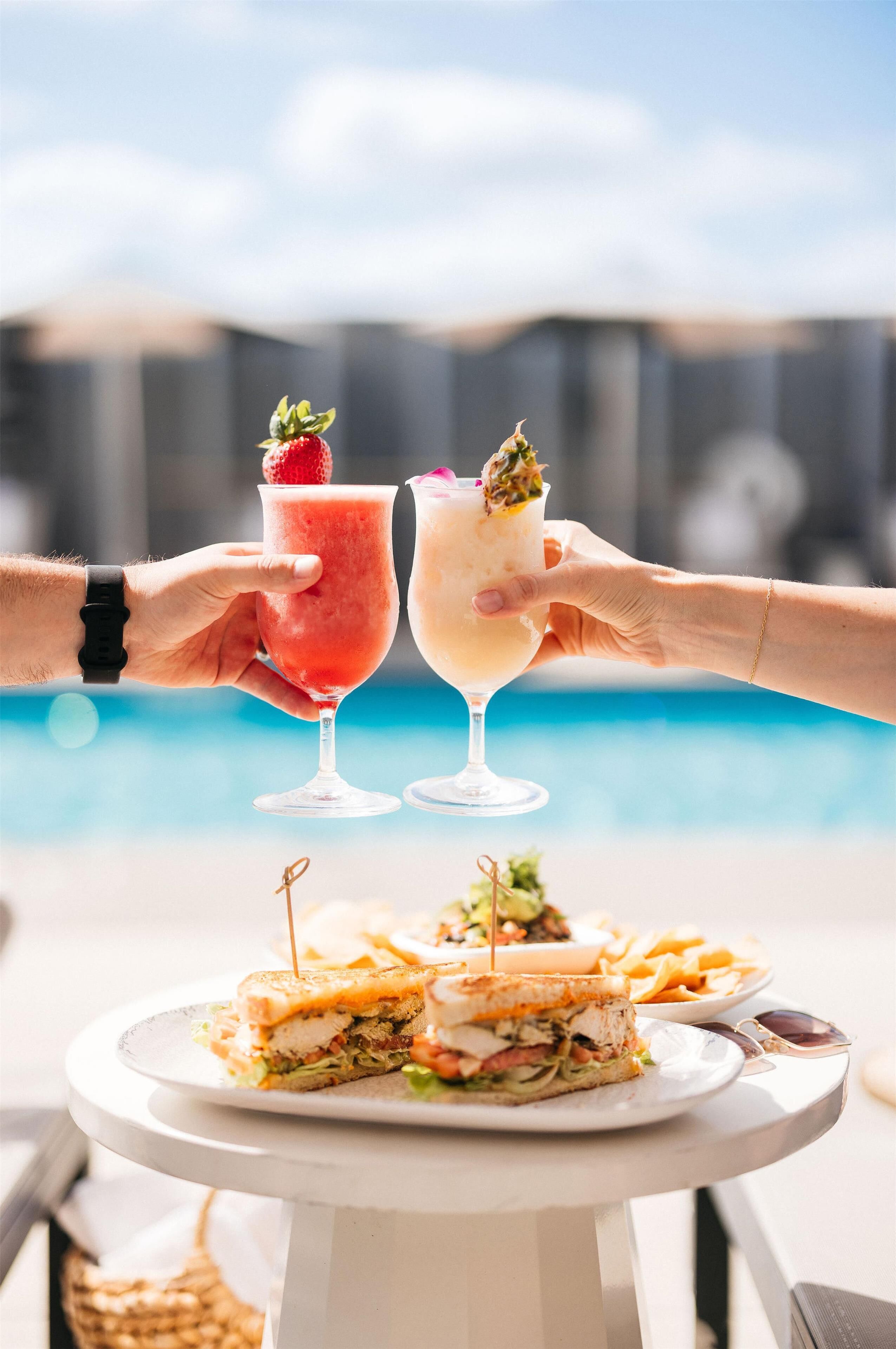 Two hands holding cocktail glasses with fruit garnishes, a plate with a sandwich, and a pool in the background.