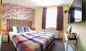 Una habitación de hotel con una cama grande, una mesita de noche, un escritorio con hervidor, un radiador y una ventana con cortinas.