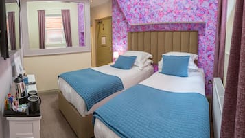 A hotel room with two beds, each with a blue quilted bedspread and matching pillows. There is a small table with a kettle and cups, and a mirror on the wall.
