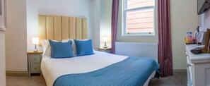 Una cama bien tendida con ropa de cama azul, una mesita de noche con una lámpara y una ventana con cortinas.