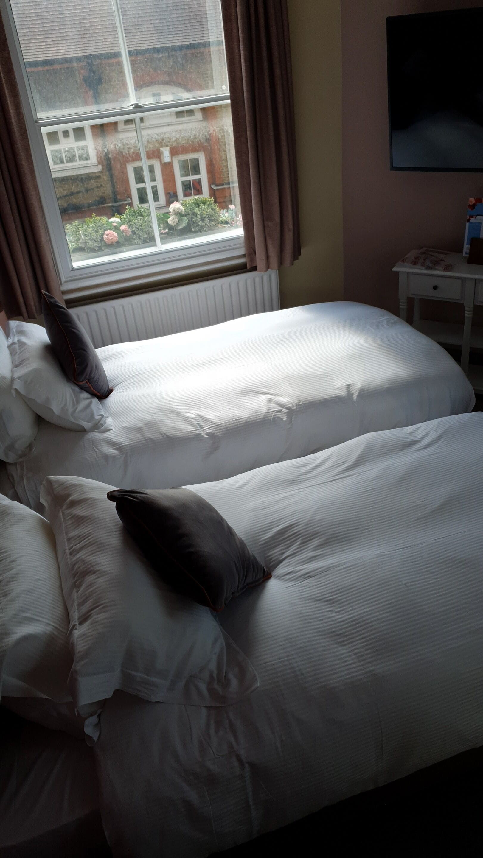 A bedroom with two beds, a window with curtains, a radiator, and a TV on the wall.