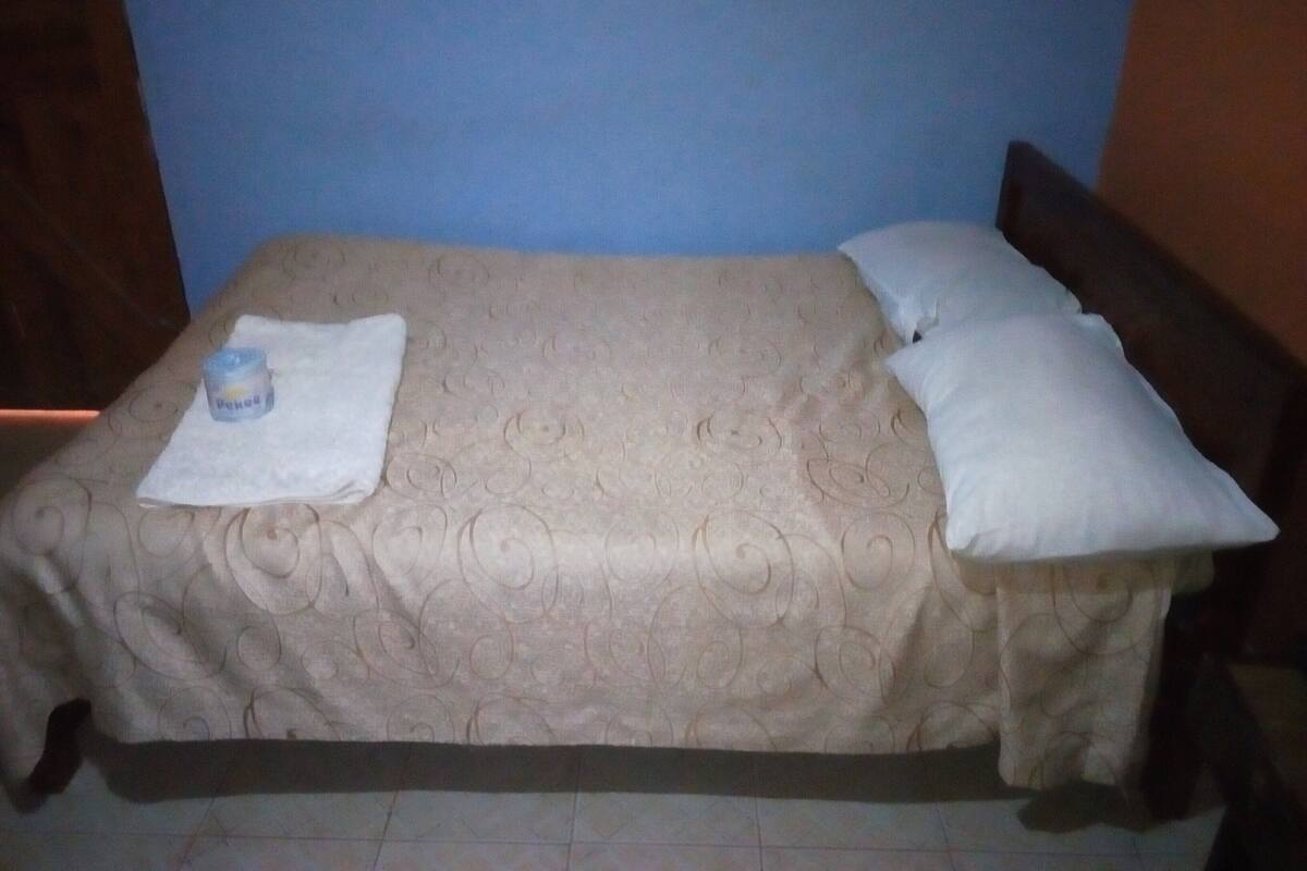 A bed with a beige patterned bedspread, two white pillows, and a small container on a white cloth.