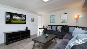 A modern living room with a flat-screen TV, a dark wooden coffee table, a grey sofa with patterned cushions, and a small side table. - Spacious Remodel-Great Freeway access-Sleeps 12 (Tucson)