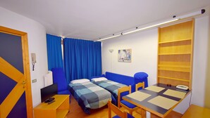 Una habitación compacta con dos camas, un escritorio con computadora, una silla y una mesita.