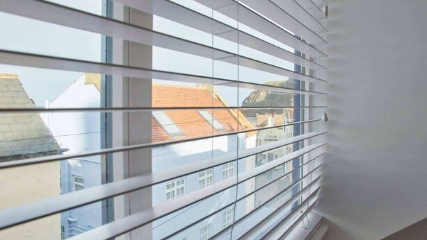 Una ventana con persianas horizontales, vista a edificios con tejados rojos y una pared blanca.