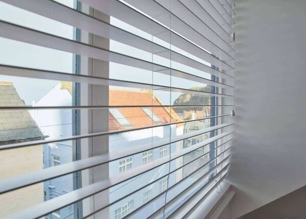 A window with horizontal blinds, a view of buildings with red roofs, and a white wall.