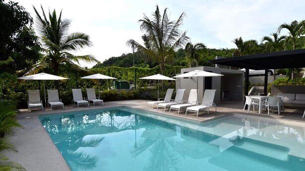 A swimming pool with white lounge chairs and umbrellas, surrounded by greenery and a covered seating area. - Agaves 203 (Santa María Huatulco)
