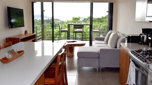 A modern kitchen with a large island, a dining area with chairs, a sofa, and a view of greenery through large windows. - Agaves 203 (Santa María Huatulco)