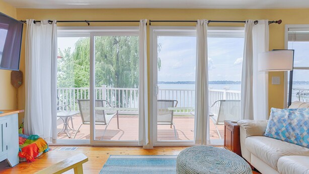 Un salon comprenant un canapé, une table basse et un téléviseur. Une porte-fenêtre coulissante donne sur une terrasse extérieure d’où l’on peut admirer un plan d’eau et des arbres.