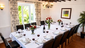 Une salle à manger avec une longue table dressée pour un repas, entourée de chaises, avec un lustre et une fenêtre ornée de rideaux.