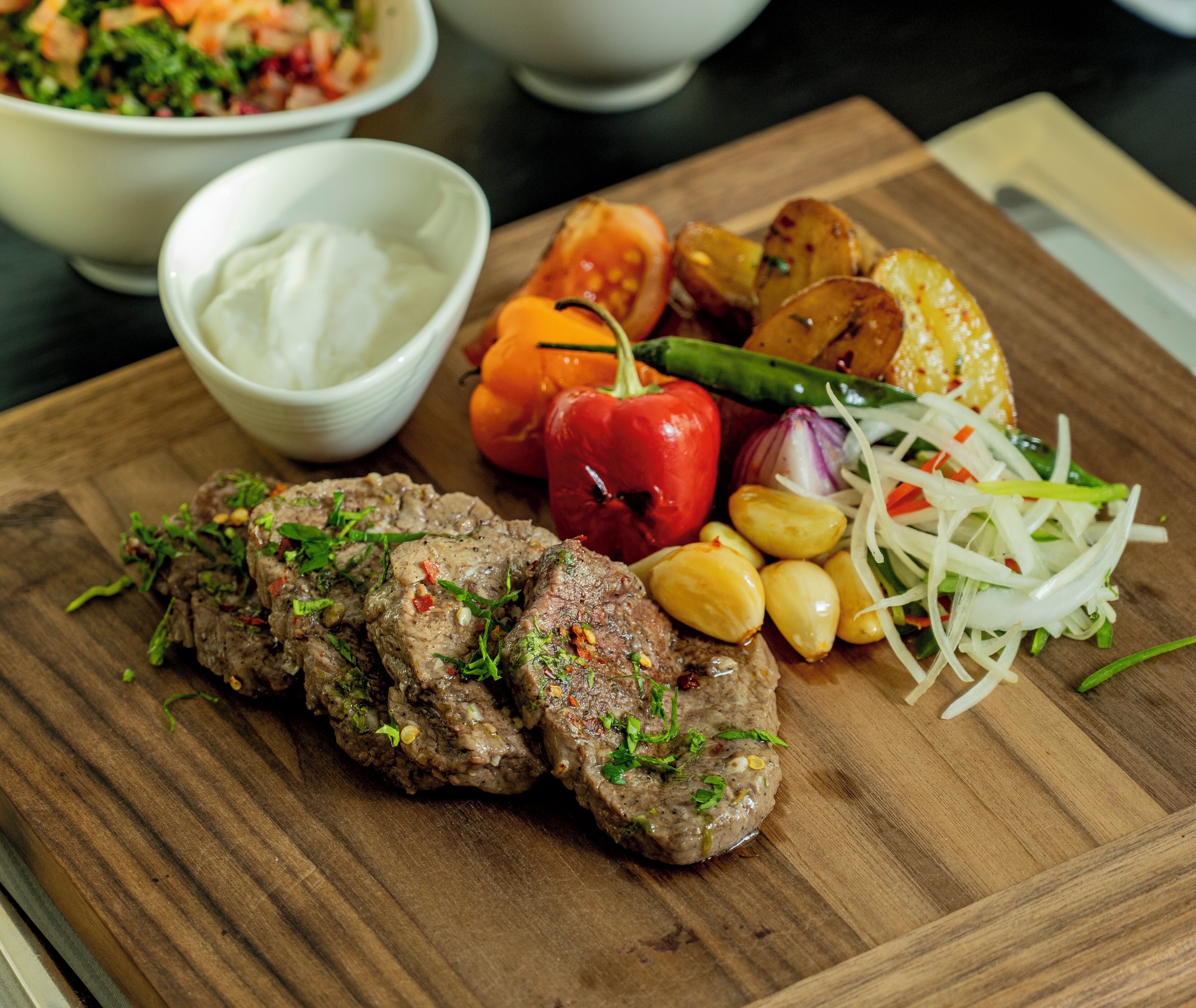 Una tabla de madera con carne a la parrilla, patatas asadas, un pimiento rojo y una ensalada.