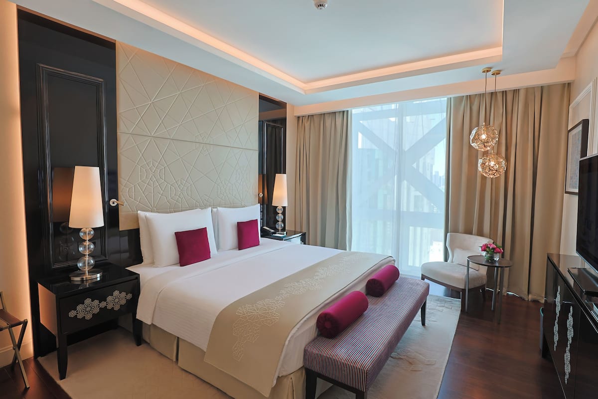 A hotel room with a large bed, bedside tables, a bench, a TV, and a view of the outside.