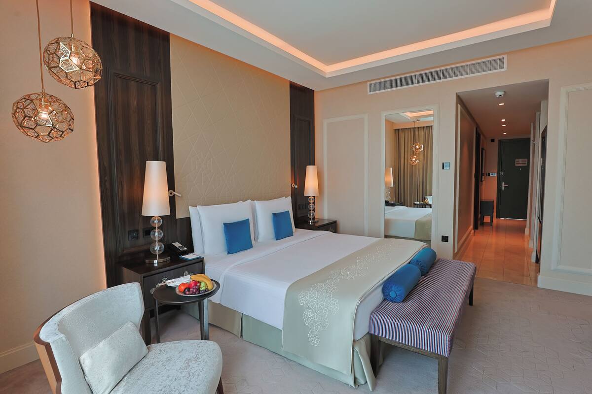 A modern hotel room with a large bed, bedside tables, a chair, and a small table with fruit.