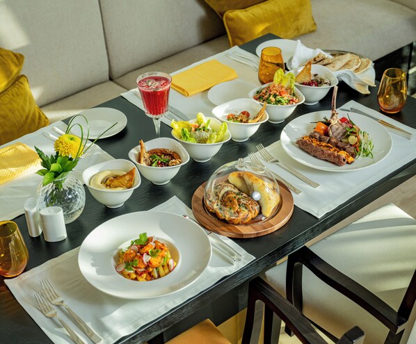 A dining table set with various dishes, including salads, meats, and bread, in a well-lit room with a sofa and cushions in the background. - Shaza Doha (Doha)